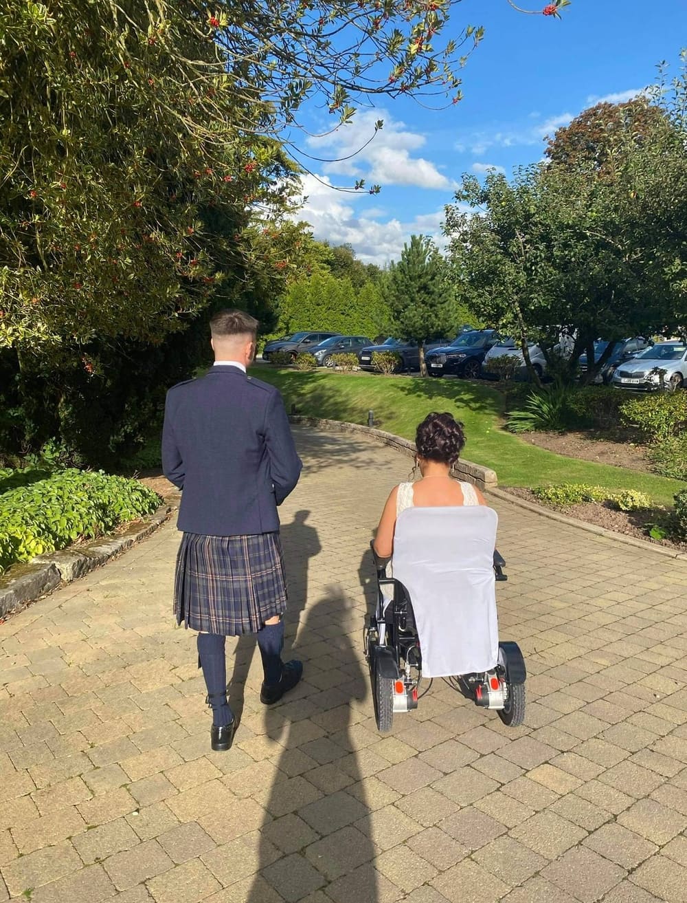 Derek, in a kilt, walking beside his Mum in her wheelchair